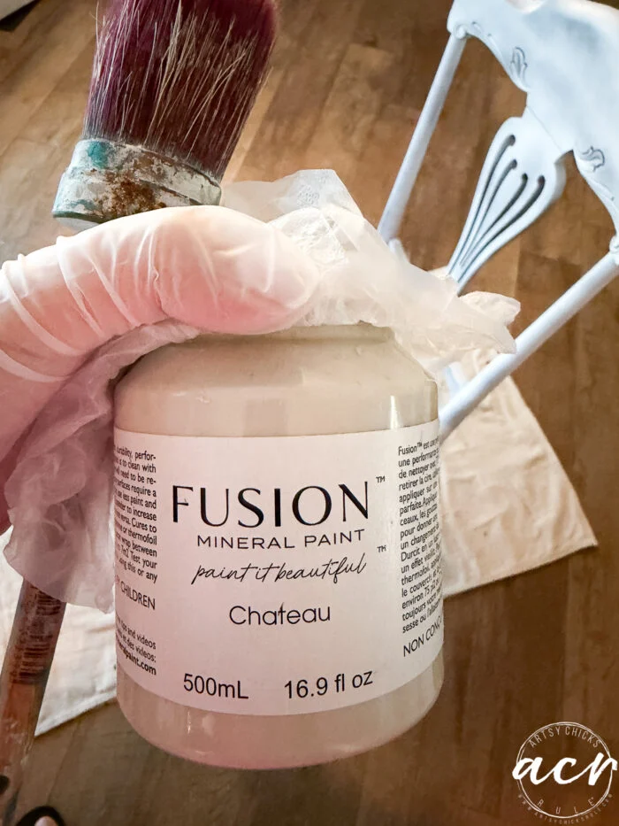 a gloved hand holds a jar of fusion mineral paint in chateau with a paintbrush, beside a partially painted blue and white vintage chair on a wooden floor—perfect for your next vintage furniture update.