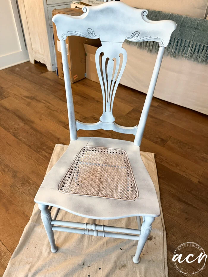 a vintage chair with a cane seat is partially painted in light blue and white, sitting on a drop cloth on a hardwood floor, with a sofa and blanket in the background—perfect for your next chair update.