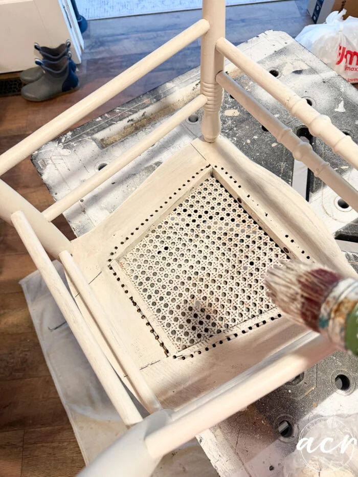 a close-up of an upside-down vintage chair being painted white, focusing on the woven cane seat. a paintbrush is visible in the lower right corner, updating this charming piece of vintage furniture with a fresh coat of paint.