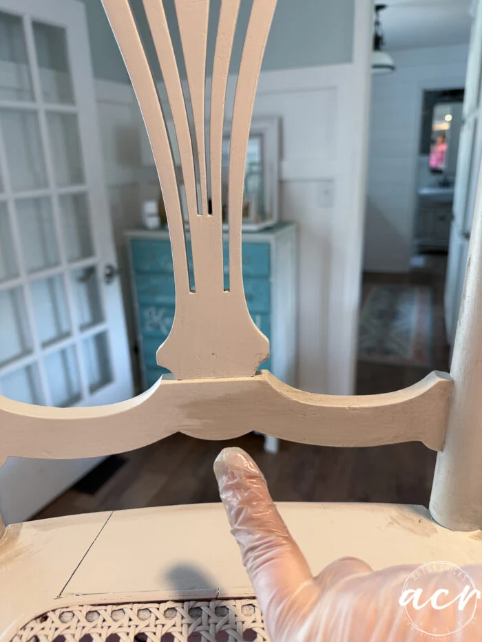 a gloved finger points at a detail on the backrest of a partially painted vintage chair, with a light blue cabinet and glass-paneled door visible in the background—a charming chair update in progress.