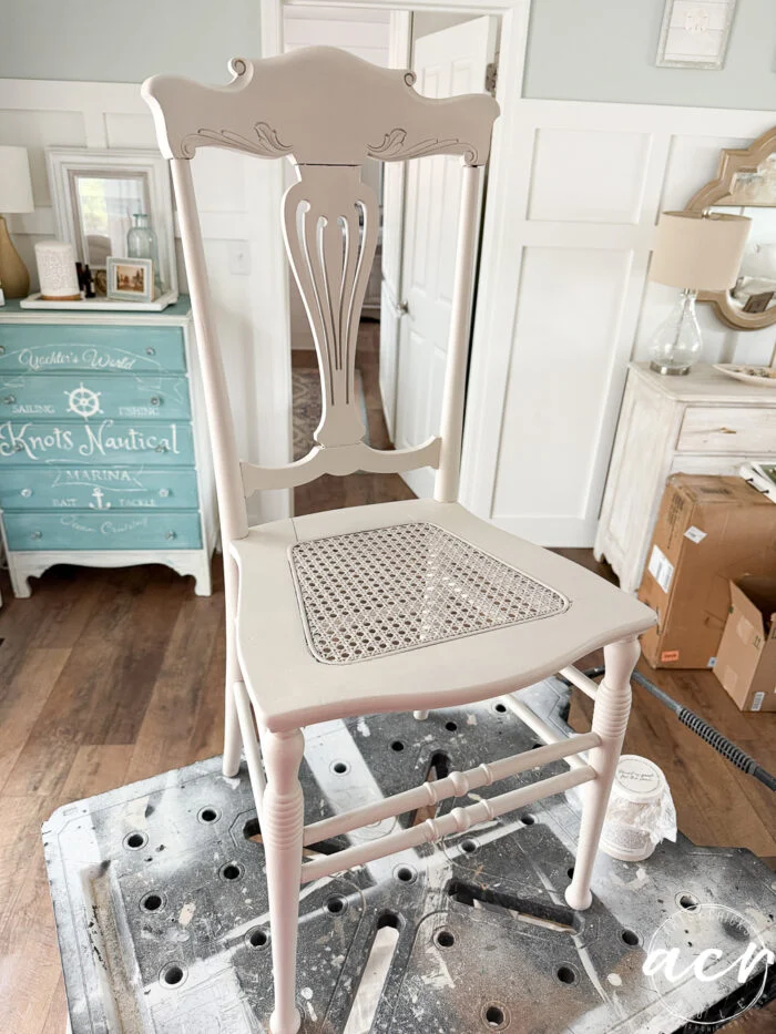 a vintage chair with a pale beige paint finish and a cane seat sits on a workbench in a bright, airy room with cottage-style decor—the perfect setting for your next charming vintage furniture update.