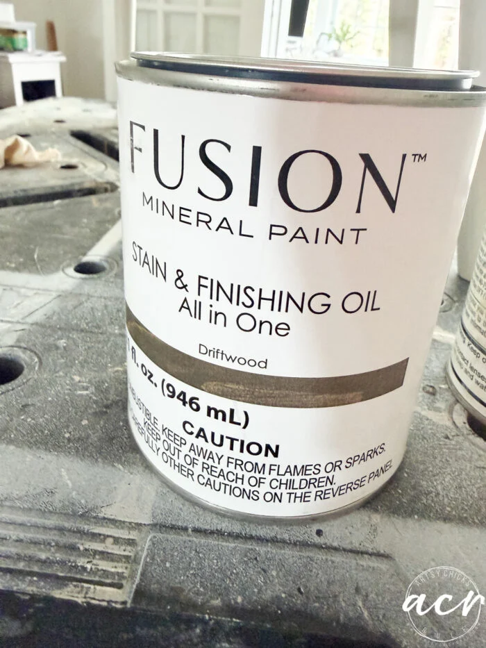 a can of fusion mineral paint stain & finishing oil (driftwood) sits on a work surface, ready for your next vintage chair update, showing the label with product details and safety cautions.