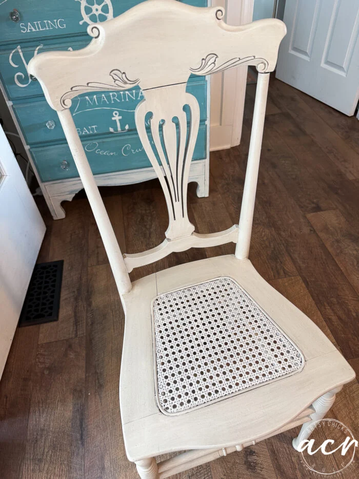 a vintage chair painted off-white with a cane seat sits on a wood floor near a blue nautical-themed dresser decorated with white marine motifs. this piece adds timeless charm to any vintage furniture collection.