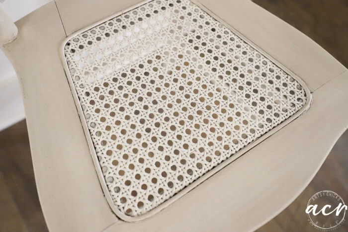 close-up of a vintage chair seat with a woven cane pattern, painted in light beige, on a dark wooden floor. the seat features a square inset and detailed lattice design, perfect for those who love vintage furniture accents.