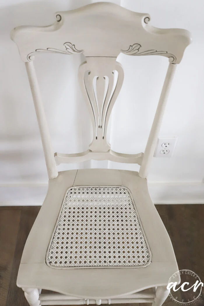 a vintage chair with ornate carvings on the backrest and a cane seat sits against a white wall and dark wood floor. an electrical outlet is visible on the wall behind this timeless piece.