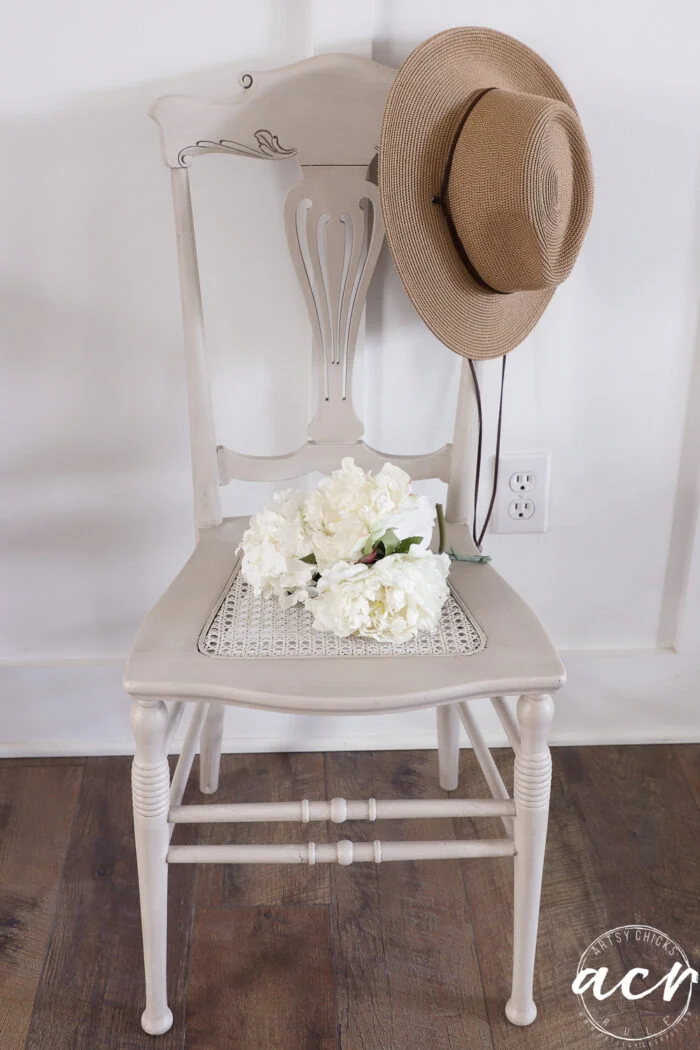 a beige vintage chair holds a straw hat draped over its back and a bouquet of white flowers on its seat, showcasing the timeless charm of vintage furniture against a white wall and wood floor.