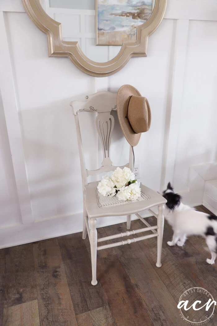 a beige vintage chair with a straw hat on the backrest and a tray of white flowers on the seat sits against a white paneled wall; nearby, a small black and white dog stands on the wooden floor—a charming scene for any vintage chair update.