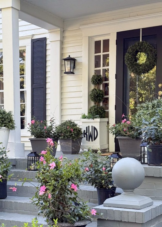 front porch of a white house decorated with potted plants and pink flowers, perfect for inspiration on how to decorate a porch for summer. black lanterns line the steps, while a green wreath adorns the door framed by white columns and dark shutters.