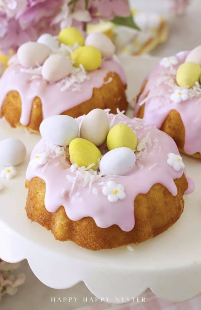mini bundt cakes with pink icing, decorated with pastel candy eggs, white edible flowers, and coconut flakes, displayed on a white cake stand.