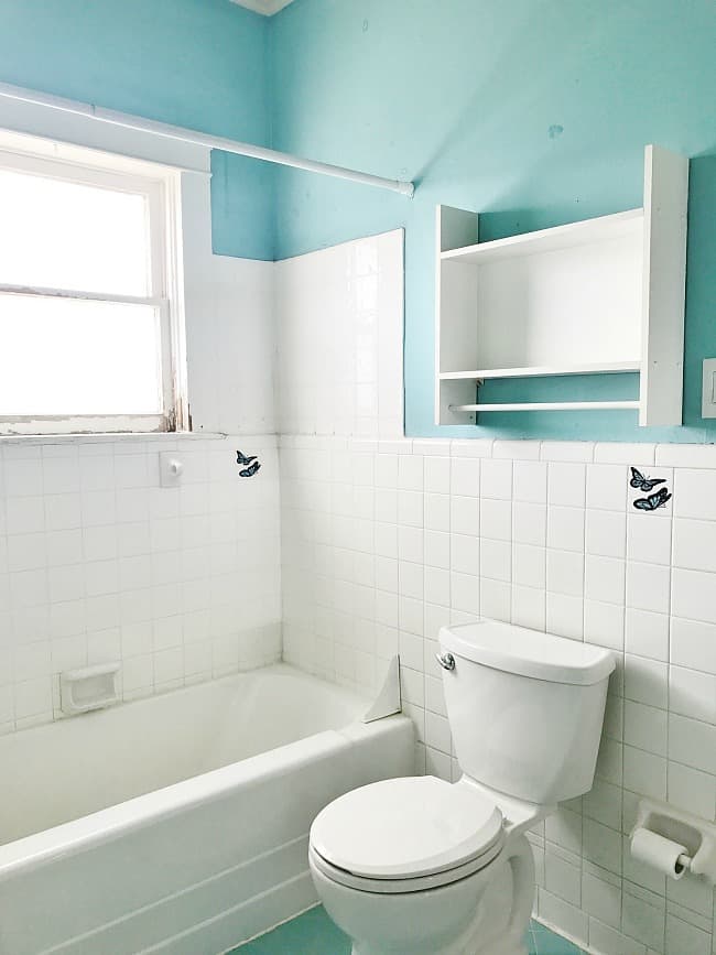 a brightly lit bathroom with white tiles and a light blue wall. it includes a bathtub, a toilet, and open shelving with small butterfly decorations on the tiles.