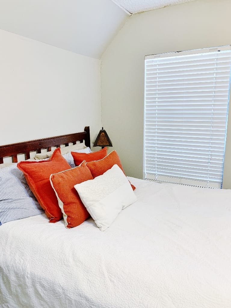 a neatly made bed with a wooden headboard and white bedding, adorned with several pillows in orange and white, located beside a window with closed white blinds.
