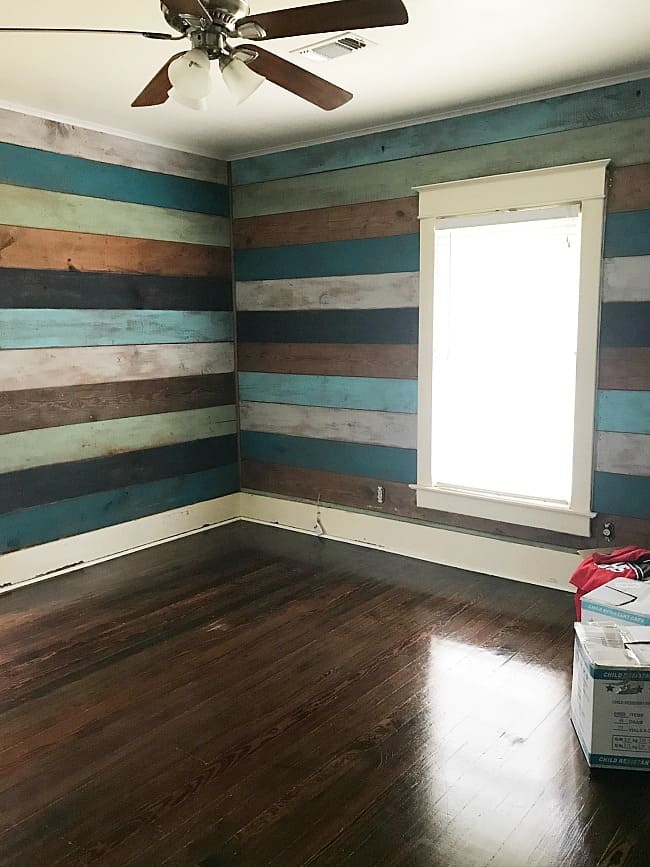 a room with dark hardwood floors and a striped accent wall in shades of blue, brown, and beige. there is a ceiling fan and a window that brightens the room.