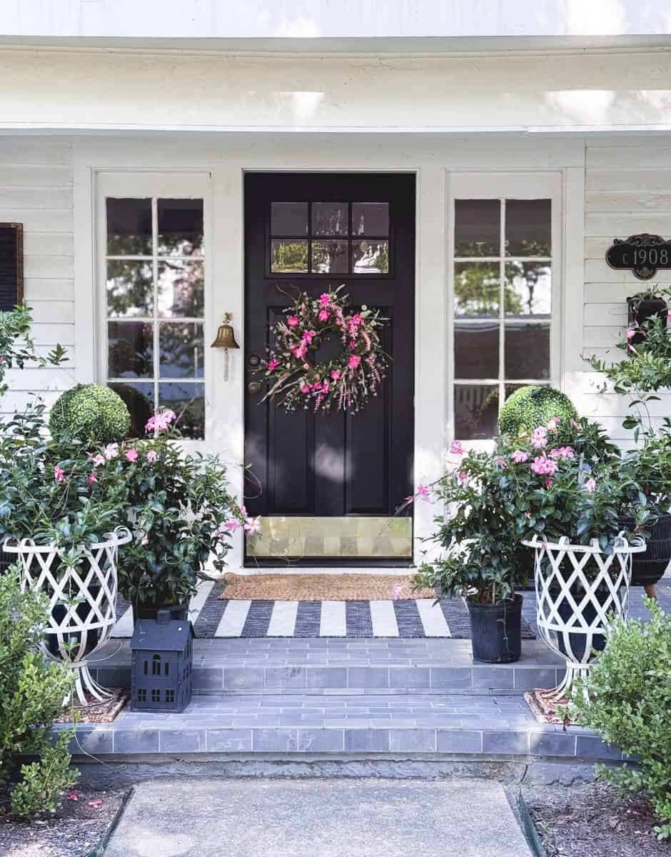 a black front door with a pink floral wreath is framed by two side windows. potted plants with pink flowers and topiary bushes are on either side of the steps, leading to a tiled porch with a striped doormat.