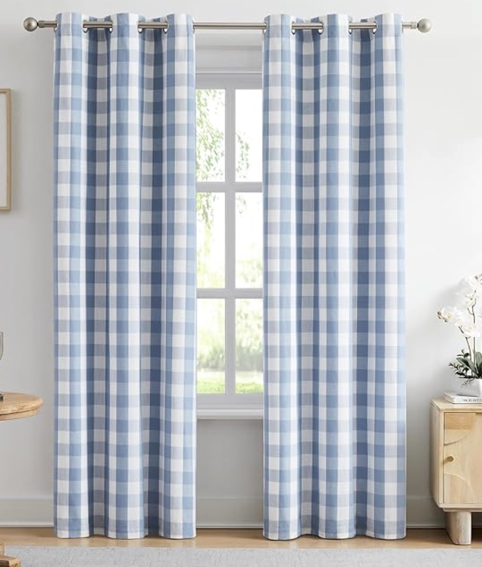 blue and white checkered curtains hang on a silver rod in front of a window, with sunlight shining through. the room has light-colored walls, a wooden table, and a small cabinet with a white flower arrangement.