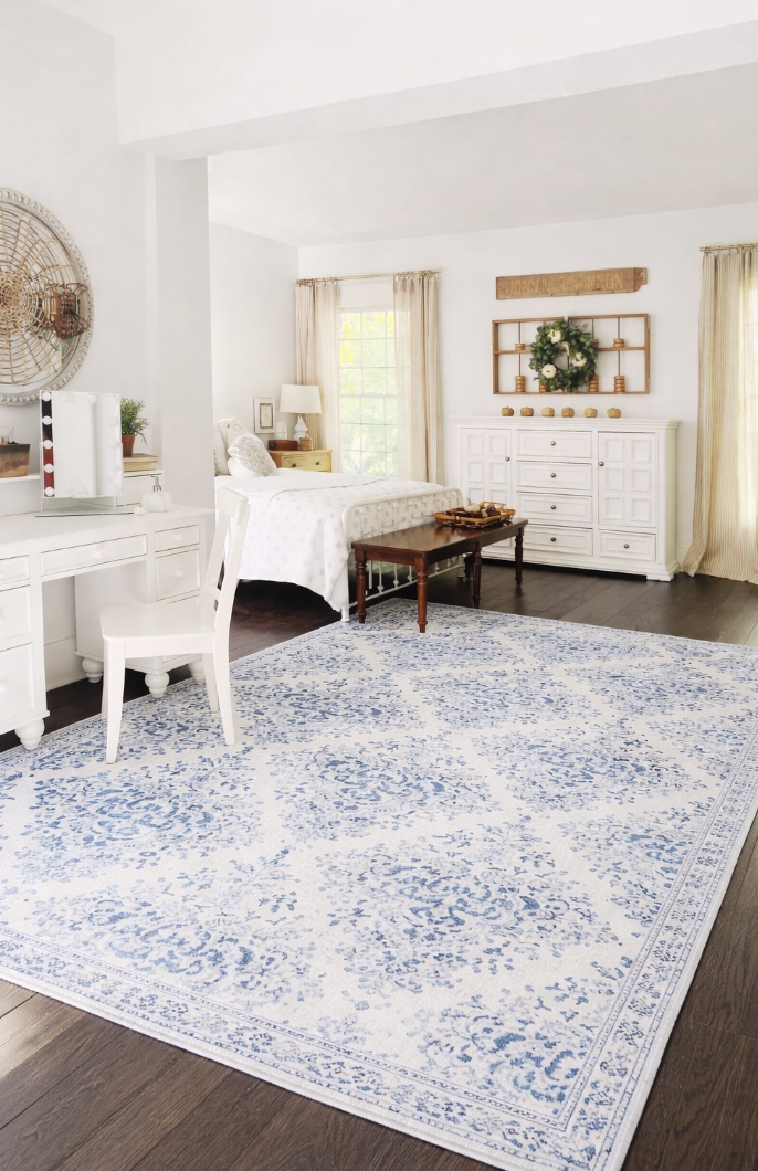 a bright, airy bedroom with white furniture, a blue and white patterned rug, a bed with white bedding, and sunlight streaming through large windows with beige curtains. a small bench sits at the foot of the bed.