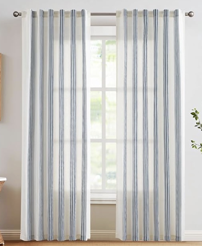 white curtains with vertical blue stripes hang on a rod in front of a large window, letting in natural light. the room has light-colored walls and a wooden floor, with a plant partially visible on the right.