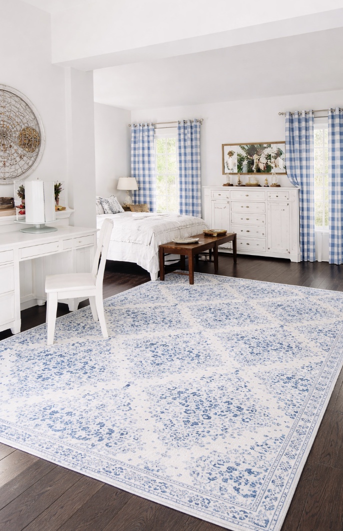 bright bedroom with white furniture, a blue-and-white floral rug, a bed with white bedding, a white chair at a vanity, blue gingham curtains, and wooden floors. natural light fills the room.