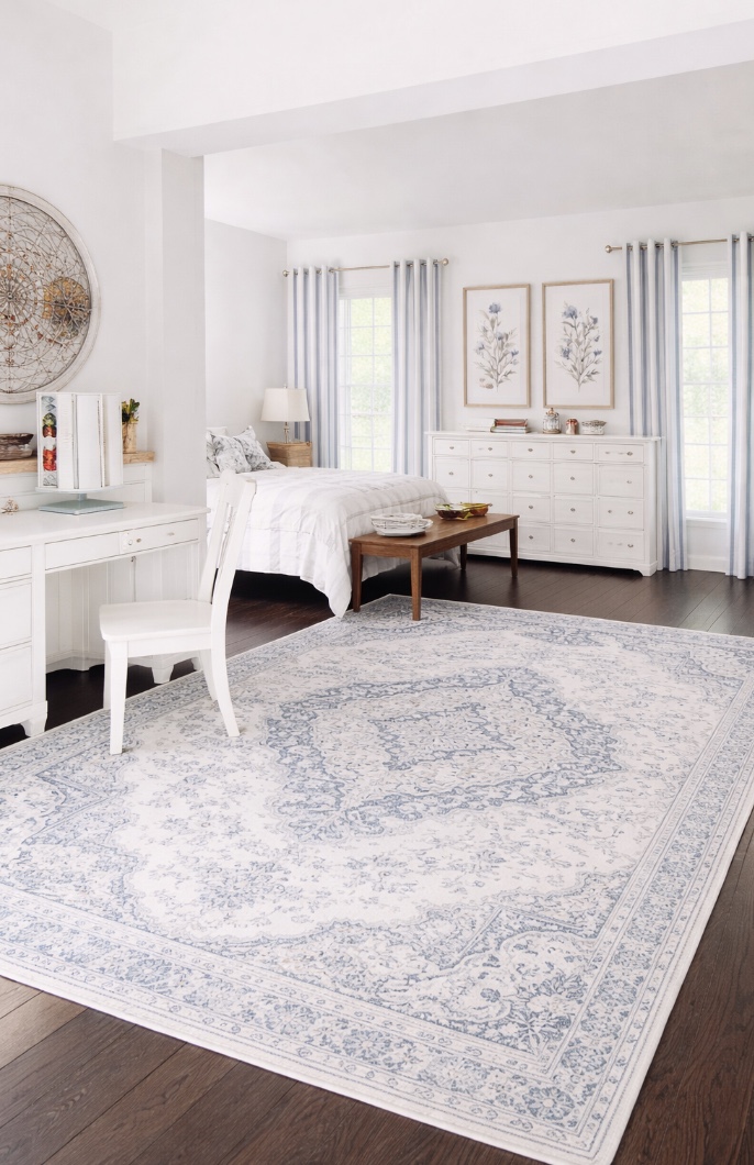 bright, airy bedroom with white furniture, a large bed, light blue and white patterned rug, wall art featuring flowers, and large windows with sheer curtains letting in natural light.