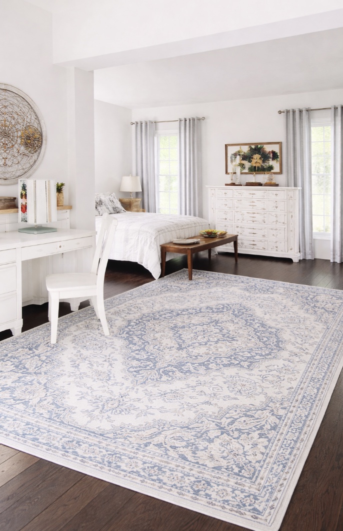 bright bedroom with white furniture, sheer curtains, dark wood floors, and a large white and blue patterned rug. artwork and decorative items are displayed on the dresser and walls, creating a cozy, elegant atmosphere.