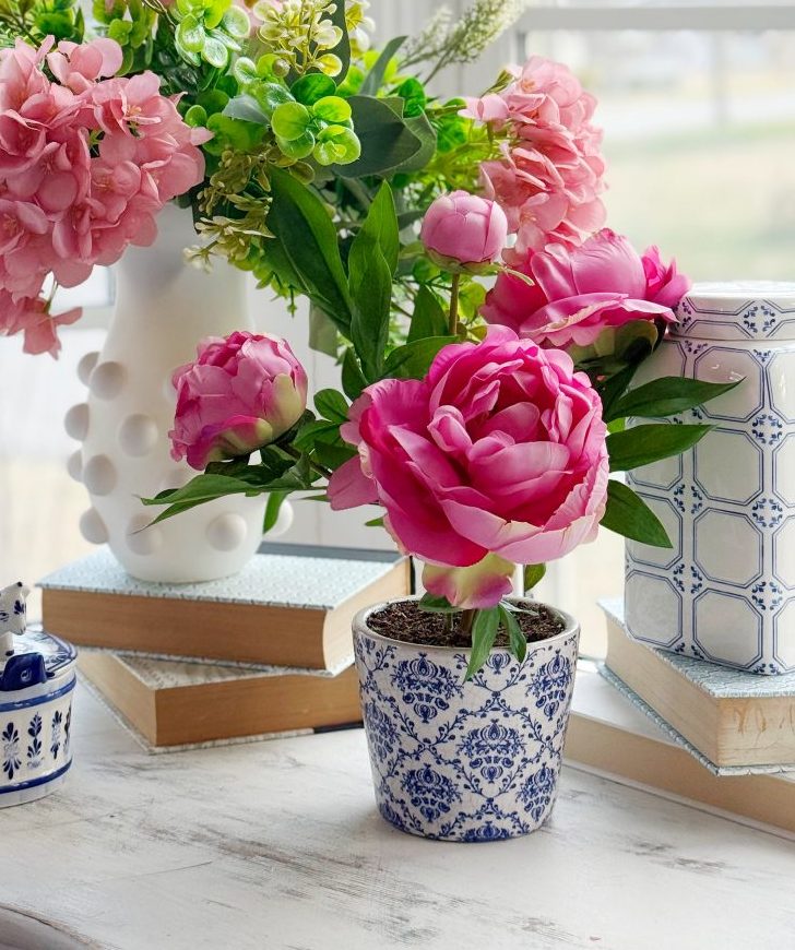 11 things i wish i bought sooner (april edition) 2 a white table by a window holds pink flowers in vases, stacked books, a porcelain box with a blue cow figurine, and a decorative jar. soft natural light brightens the cheerful floral arrangement.