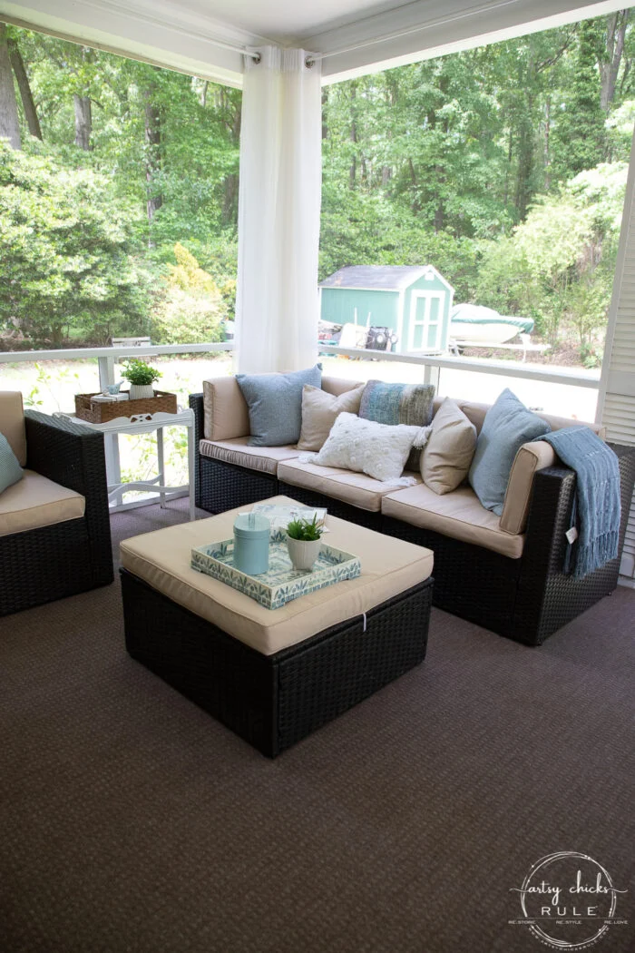 office reveal (sunroom) - artsy chicks rule® 55 a cozy screened-in porch refresh featuring wicker patio furniture, beige cushions, blue and white throw pillows, a matching ottoman, side table with decor, and views of lush green trees outside.