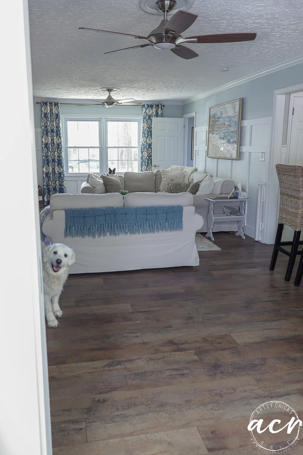 office reveal (sunroom) - artsy chicks rule® 59 a white dog peeks playfully around a doorway into a cozy living room and sunroom with a white sofa, blue blanket, patterned curtains, wooden floor, and ceiling fans.