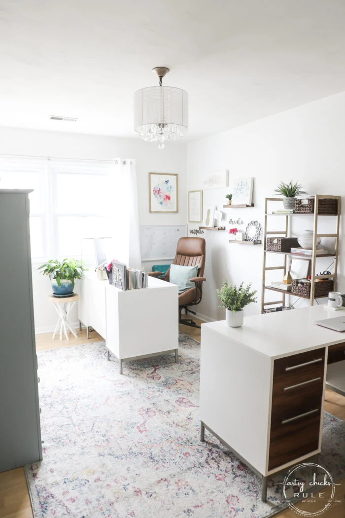 office reveal (sunroom) - artsy chicks rule® 57 bright, modern mid century modern office with white desks, a patterned rug, shelving with decor and storage, a brown chair, potted plants, wall art, and a window letting in natural light.