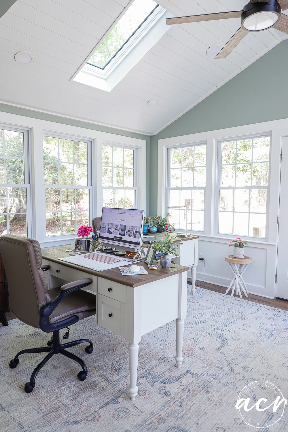 office reveal (sunroom) - artsy chicks rule® 61 a bright sunroom home office with large windows, a skylight, a white desk with a computer, brown office chair, plants, and decorative items; sunlight fills the room, creating a cozy workspace perfect for an office reveal.