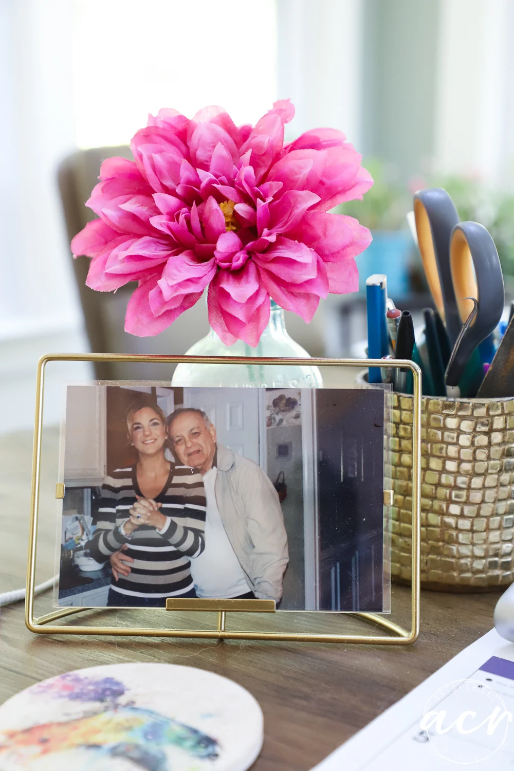 office reveal (sunroom) - artsy chicks rule® 63 a framed photo of two smiling people sits on a desk next to a vase with a large pink flower and a gold holder with pens and scissors. the cozy workspace, perfect for an office reveal, feels both welcoming and inspiring.