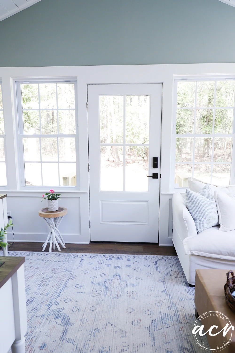 office reveal (sunroom) - artsy chicks rule® 73 a bright living room with large windows, a white door, a white sofa with pillows, and a small round side table holding a potted plant feels like a sunroom sanctuary. light patterned rug on the floor, trees visible outside.