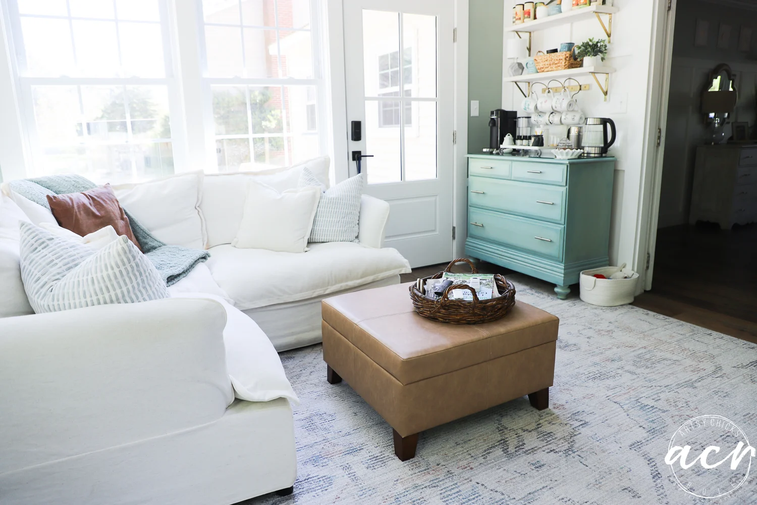 office reveal (sunroom) - artsy chicks rule® 77 bright living room doubles as a sunroom, with a white sofa, beige ottoman with wicker tray, blue sideboard with coffee supplies, open shelves with cups, and large windows. cozy, airy, and inviting decor perfect for an office reveal.