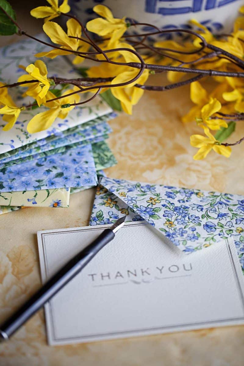 a thank you card and pen rest on a table with floral-patterned fabric squares and yellow flowers, creating a warm, inviting scene.