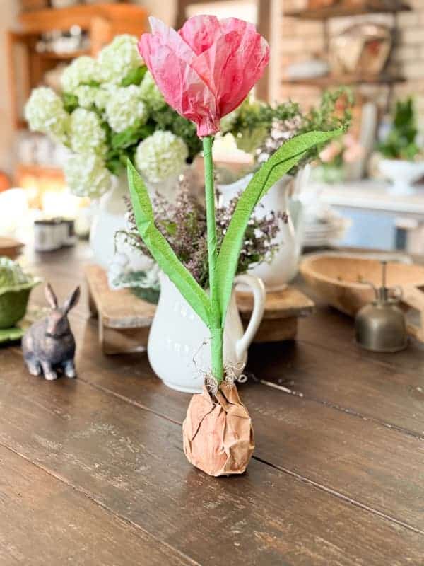 a handmade tulip craft made of pink tissue paper and green paper stands on a wooden table, with a faux brown bulb base. in the background are white pitchers with flowers and a decorative ceramic bunny.