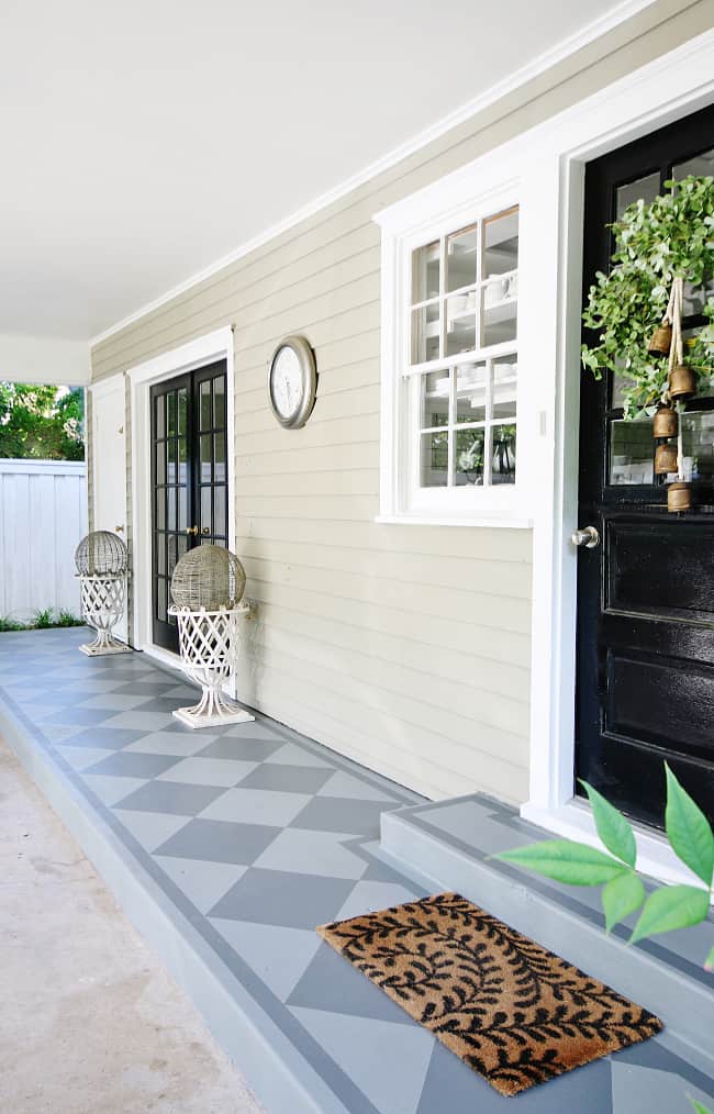 a welcoming home entrance featuring a black front door with white trim, a painted concrete patio with stylish wicker chairs, and decorative plants, conveying an inviting and trendy porch design.