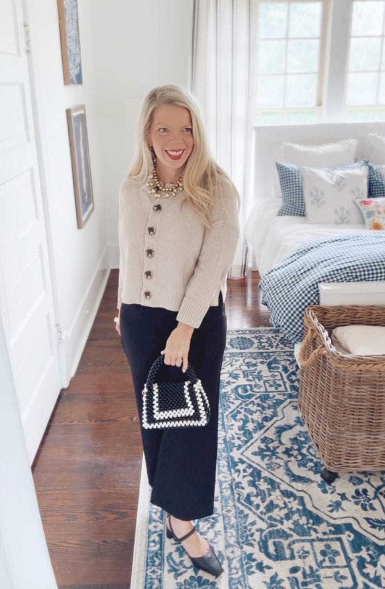 a woman with long blonde hair, wearing a beige buttoned cardigan, black wide-leg pants, and black shoes, stands in a bright bedroom holding a black and white beaded handbag. blue and white decor accents the room.