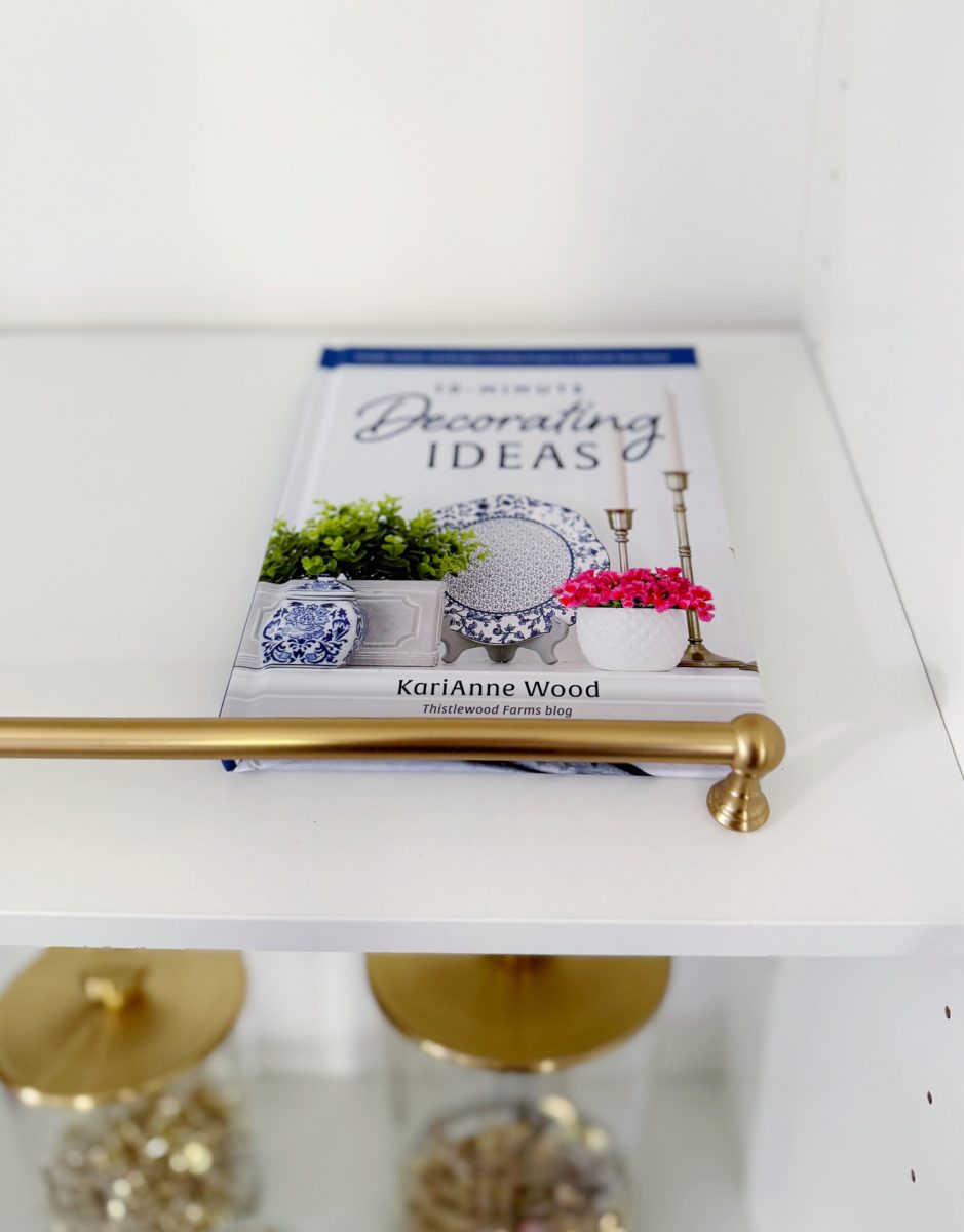 a decorating book titled "10-minute decorating ideas" by karianne wood rests on a white shelf with gold hardware. below, glass jars with gold lids contain small decorative items.