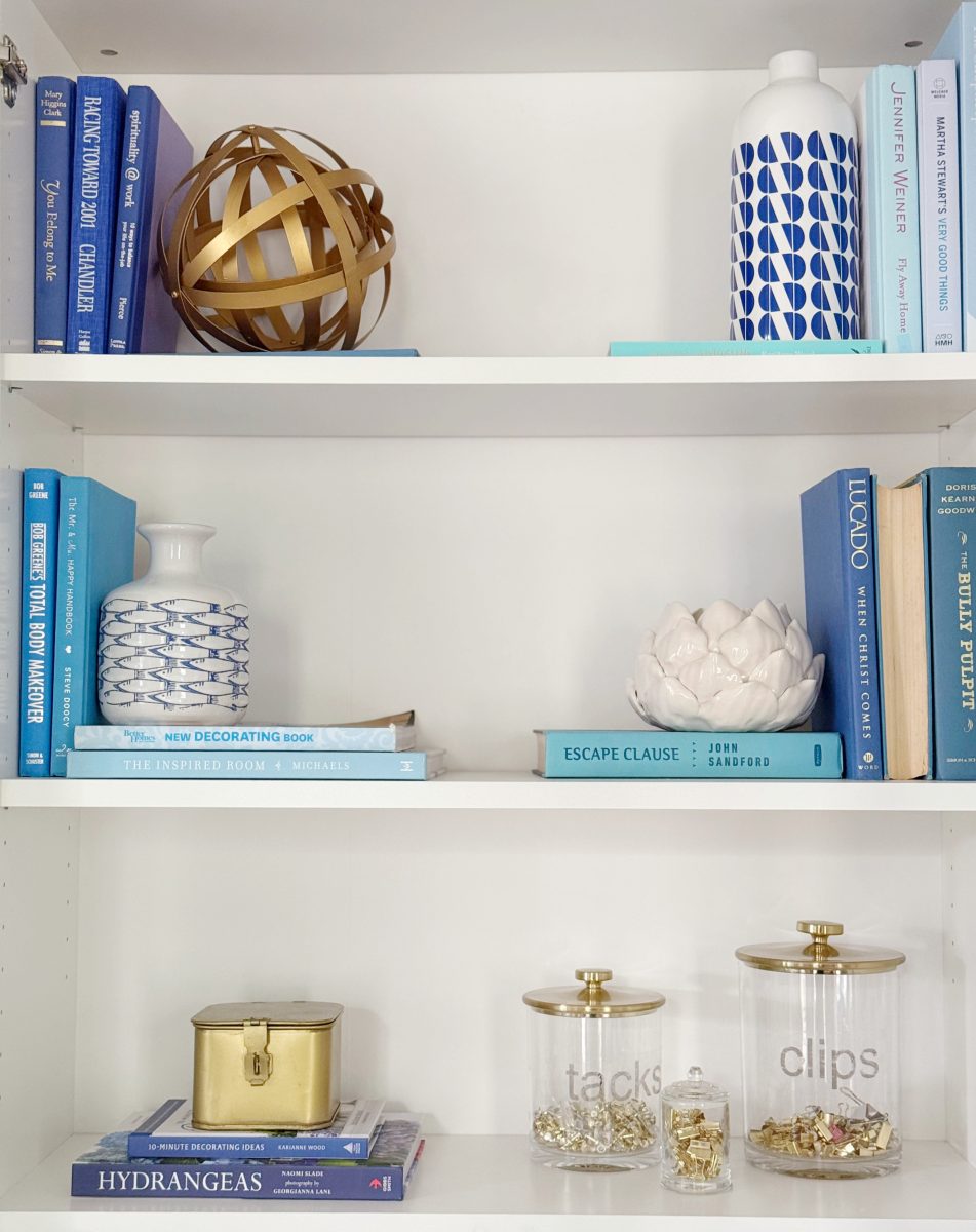 a white bookshelf displays blue and white books, a gold decorative sphere, patterned vases, glass jars labeled "tacks" and "clips" with gold lids, and a gold box, all arranged neatly on three shelves.