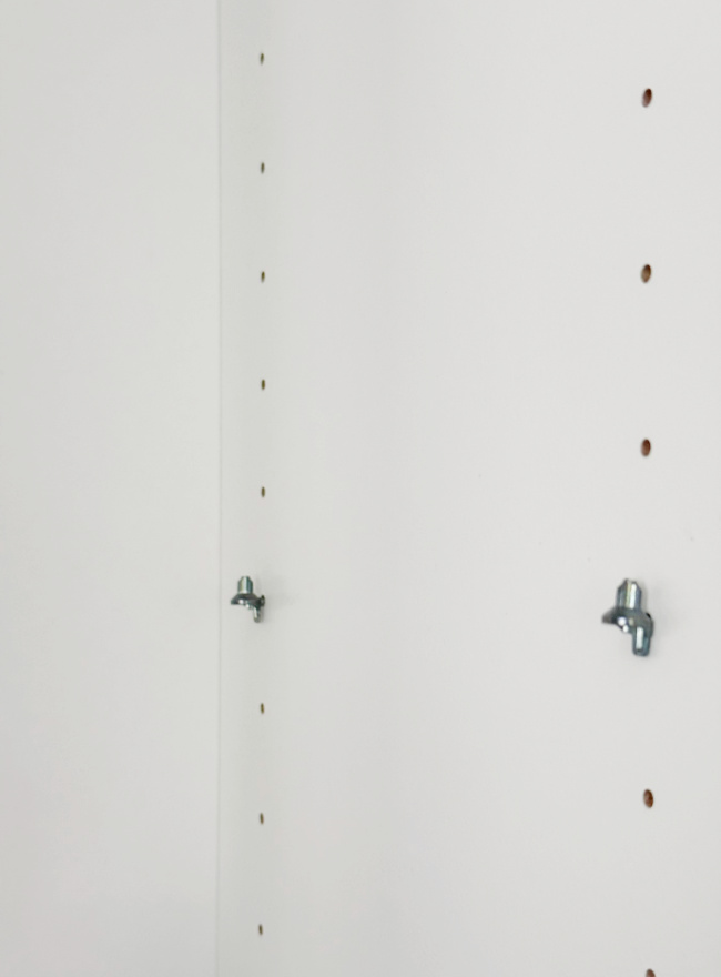 close-up of a white shelving unit side panel with evenly spaced holes and two metal shelf support pegs inserted into the holes, ready to hold a shelf.