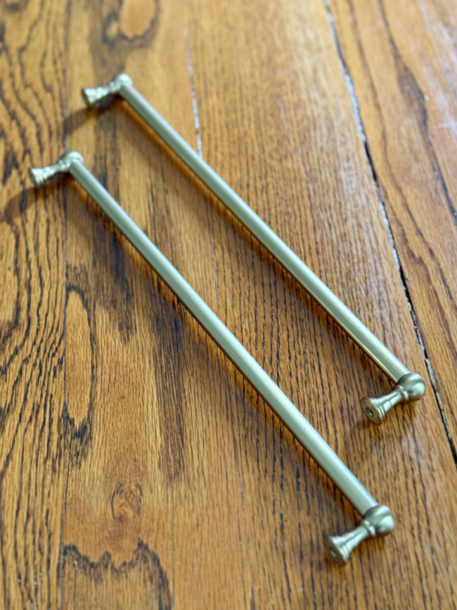 two metallic drawer or cabinet handles with decorative ends are placed parallel on a wood surface with a natural grain pattern.