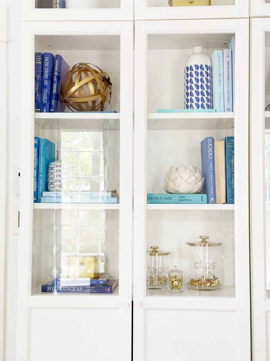 white shelves with glass doors display blue books, a decorative wooden sphere, vases, a white lotus ornament, and two glass jars labeled "tacks" and "clips" filled with small office supplies.