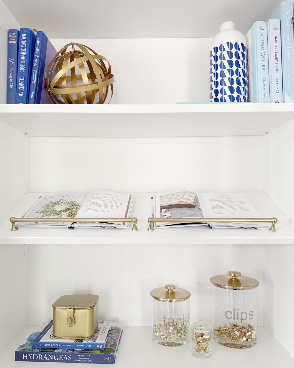 a white bookshelf with blue books, a gold decorative sphere, two open books on gold stands, a bottle with blue leaf patterns, and clear jars labeled "snacks" and "clips" filled with snacks and binder clips.
