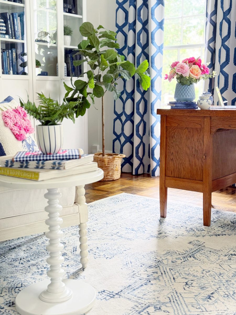 bright living room with a white side table holding books and a potted plant, blue and white patterned curtains, a large window, wooden desk with a vase of pink flowers, and a leafy plant near a white sofa.