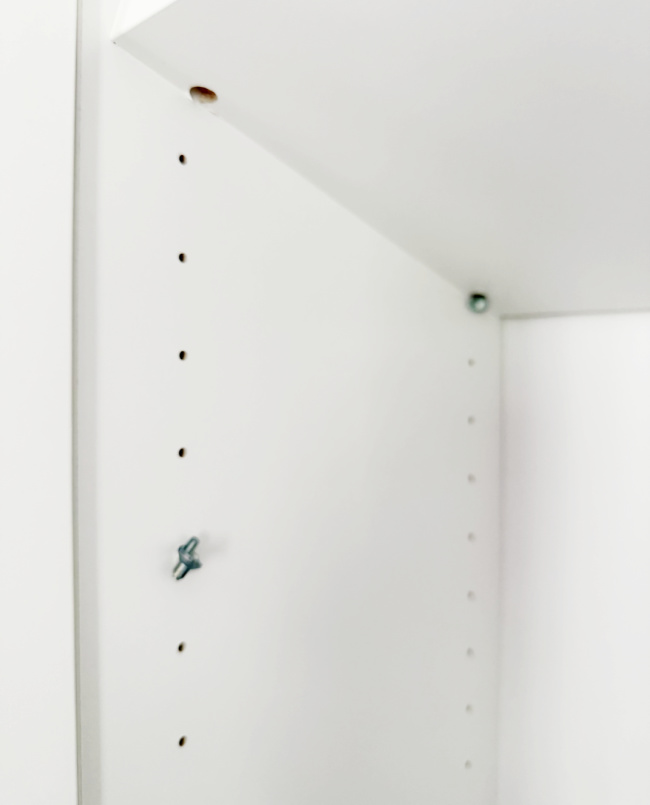 a close-up of the inside corner of a white shelving unit, showing adjustable shelf holes along the side and a screw partially inserted into one of the holes.