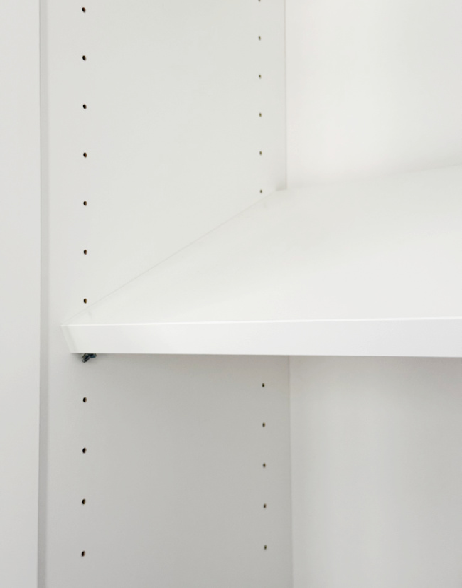 a close-up of a white shelf partially inserted into a shelving unit, with adjustable holes visible along the upright side panels.