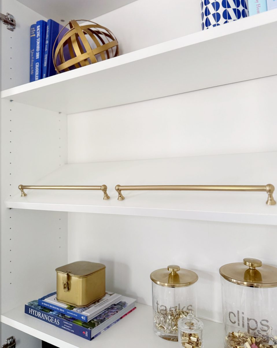 a white shelving unit with two gold rods, blue books, a gold decorative orb, a gold box, and clear jars labeled "tacks" and "clips" filled with gold office supplies.