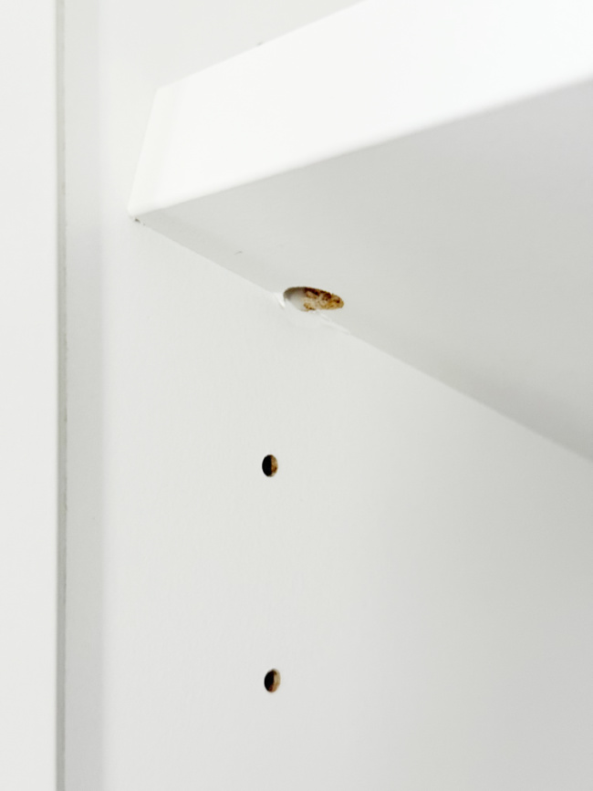 a close-up of a white shelf with two empty peg holes and a chipped corner showing exposed wood and a brownish mark.