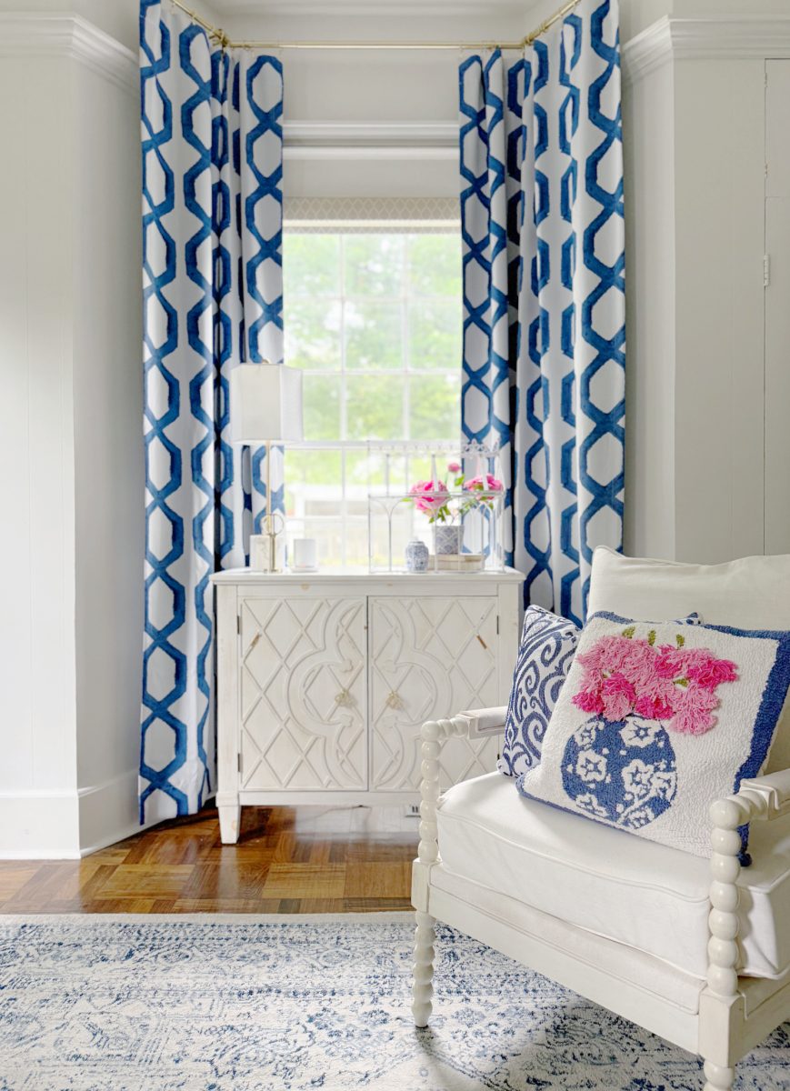 a bright, elegant room with a white armchair, blue and white patterned pillow, white cabinet, lamp, fresh flowers, blue geometric curtains, and a window letting in natural light. a blue and white rug covers the hardwood floor.