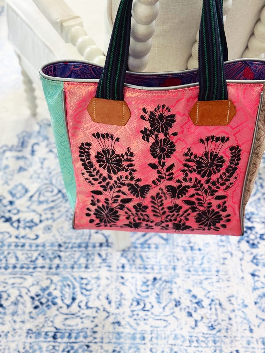 a tote bag with black floral embroidery on a pink background hangs on a white, spindle-back chair above a blue and white patterned rug. the bag has leather handles and multicolored panels.