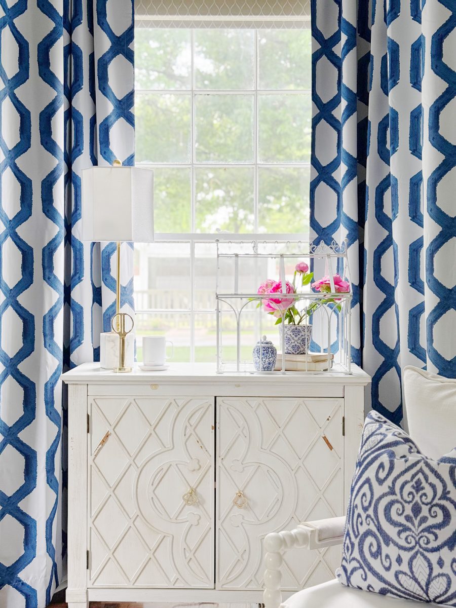 a bright room with blue and white patterned curtains, a white cabinet, a lamp, a vase with pink flowers, and decorative items. a window lets in natural light, and there is a patterned pillow on a white chair.