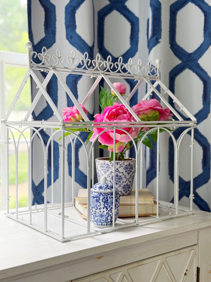 a white decorative metal greenhouse frame holds a blue and white vase with pink flowers, a small blue and white jar, and stacked books. the setup is on a white table by a window with geometric blue curtains.